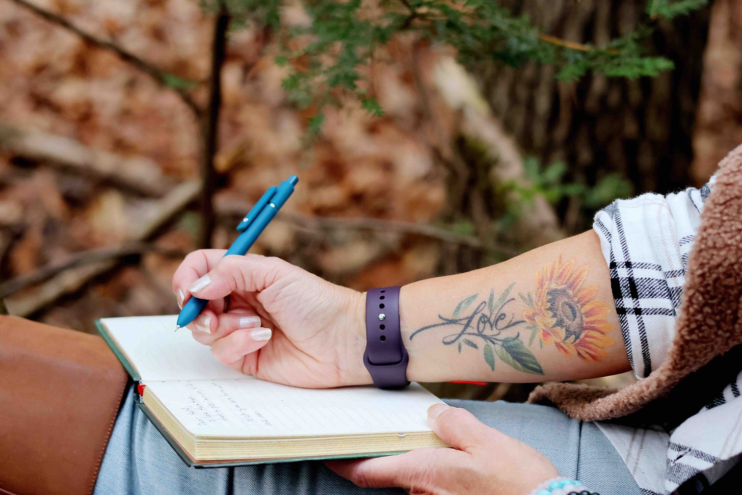 woman holding a pen over her journal. Has a tattoo of a sunflower with the word LOVE