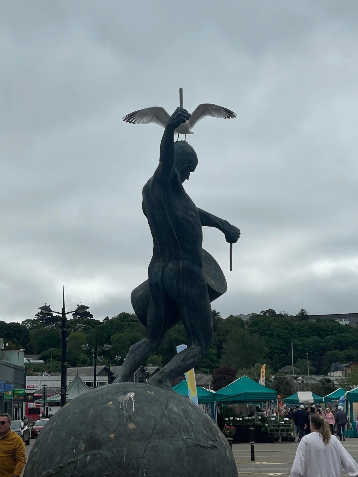 Colour photograph showing the rear view of a tall green statue of a naked man banging a drum on a ball. A gull is standing on his head and stretching its wings. In the background are shoppers and tented market stalls. Colour photograph showing the rear view of a tall green statue of a naked man banging a drum on a ball. A gull is standing on his head and stretching its wings. In the background are shoppers and tented market stalls.