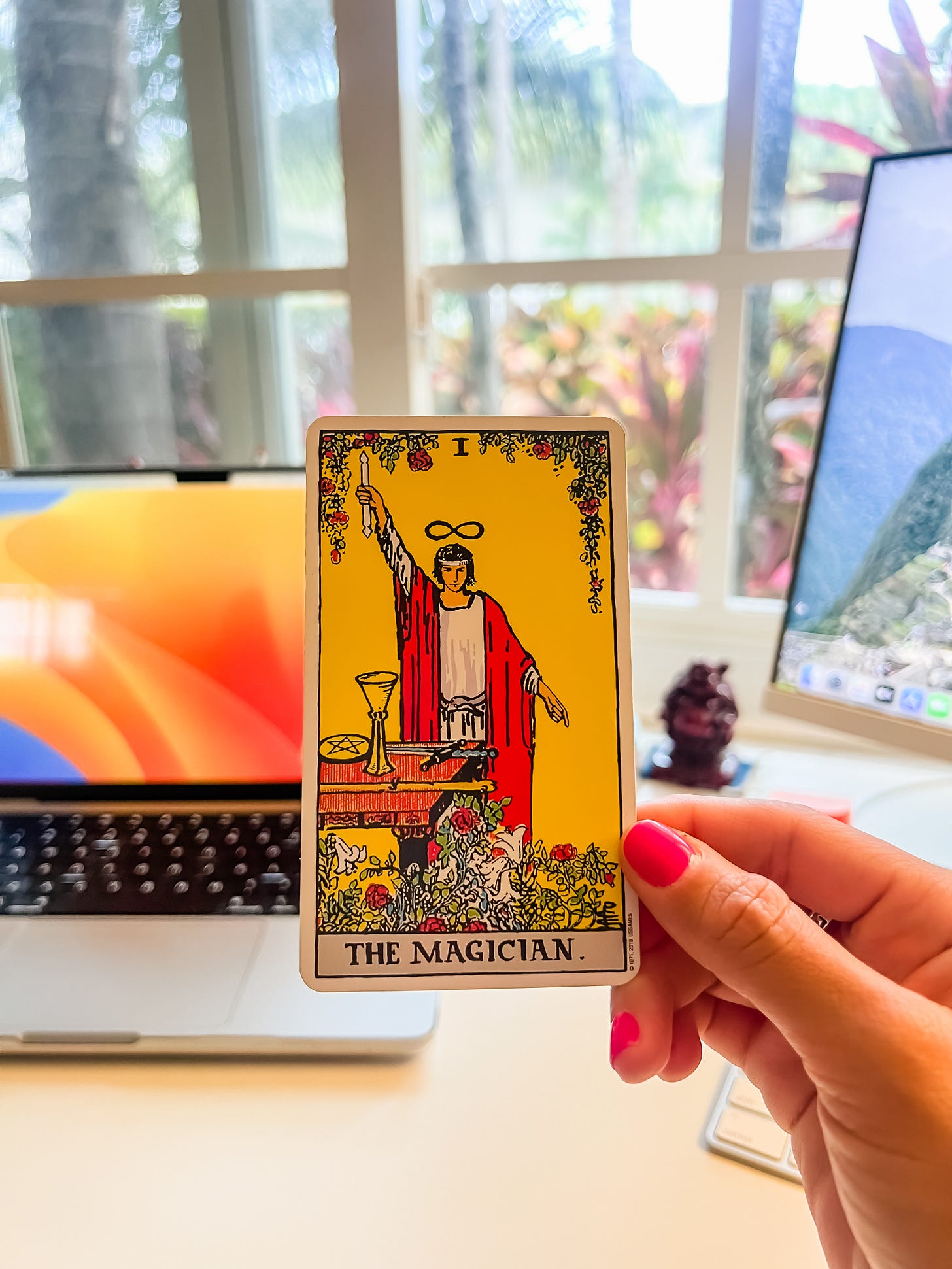 A photo of a hand holding up a tarot card, The Magician, with a laptop and computer monitor and window in the background.