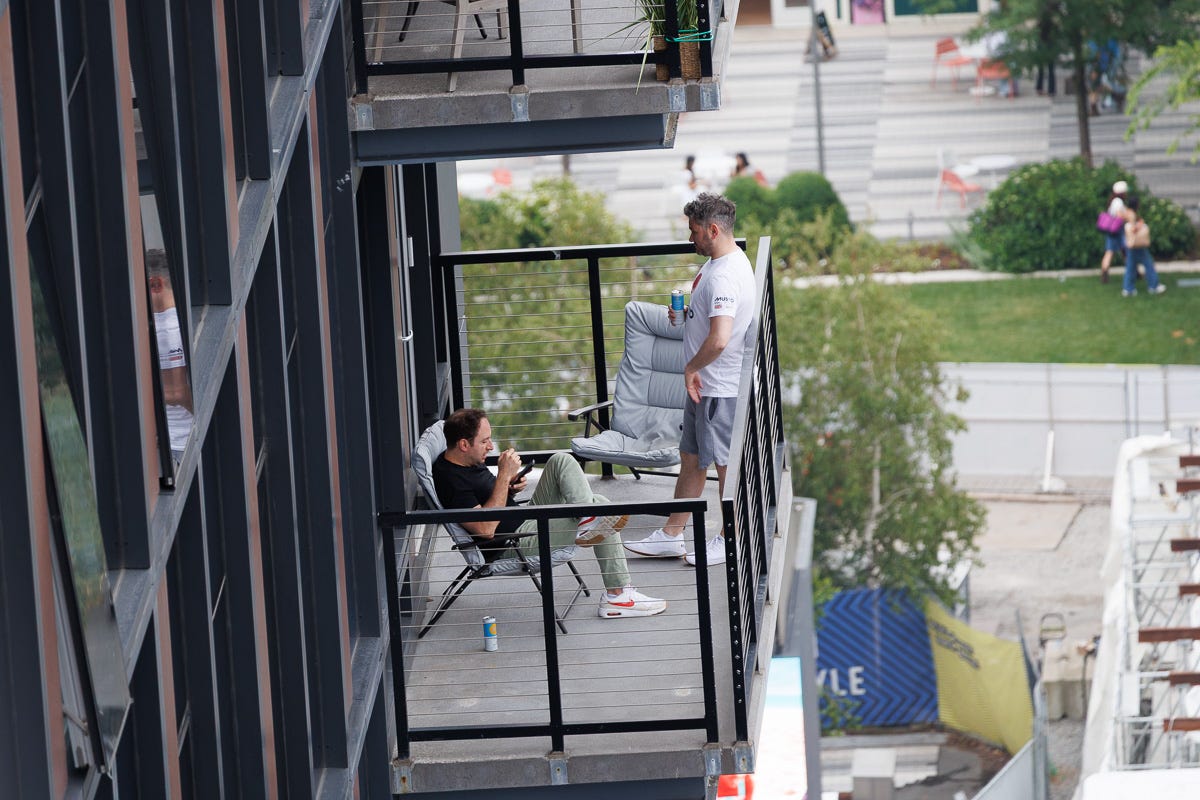 Two men chat on a modern apartment balcony, one seated and the other standing with a drink, framed against a backdrop of green space and a city plaza below. Two men chat on a modern apartment balcony, one seated and the other standing with a drink, framed against a backdrop of green space and a city plaza below.