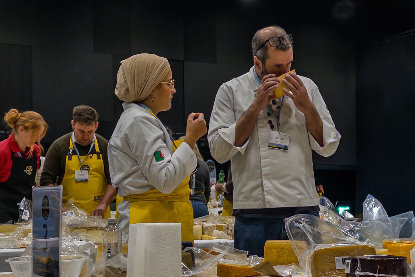 Zwei Jury-Mitglieder an den World Cheese Awards 2025 stehen an einem Tisch voller Käseproben. Die Frau links trägt ein helles Kopftuch und eine weisse Jacke, hält ein kleines Stück Käse in der Hand und spricht mit dem Mann neben ihr. Der Mann riecht konzentriert an einem grösseren Stück Käse. Im Hintergrund arbeiten weitere Personen in gelben Schürzen an ihren Verkostungstischen.