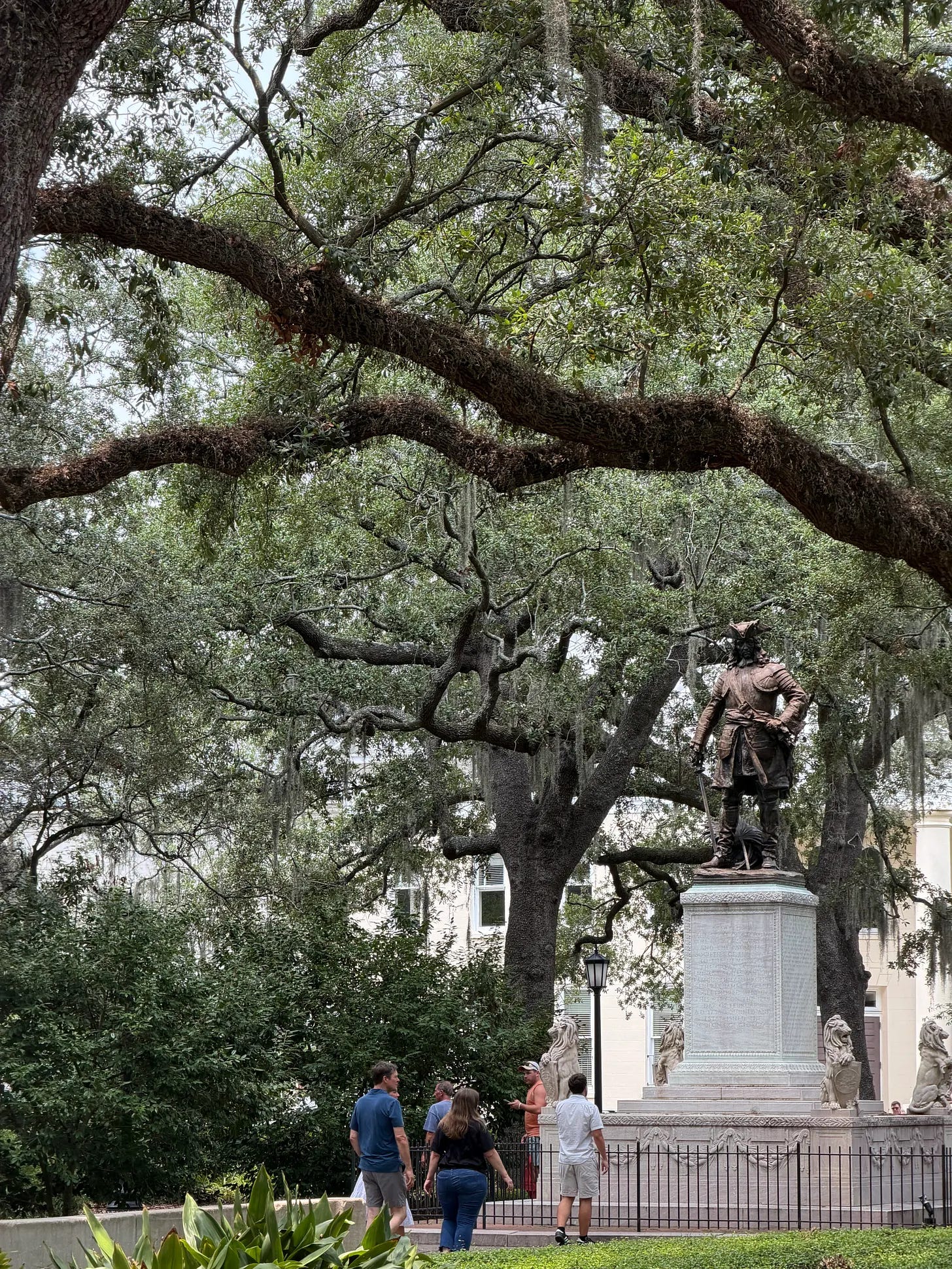People walk through a park featuring a large statue, and oak trees covered in moss.