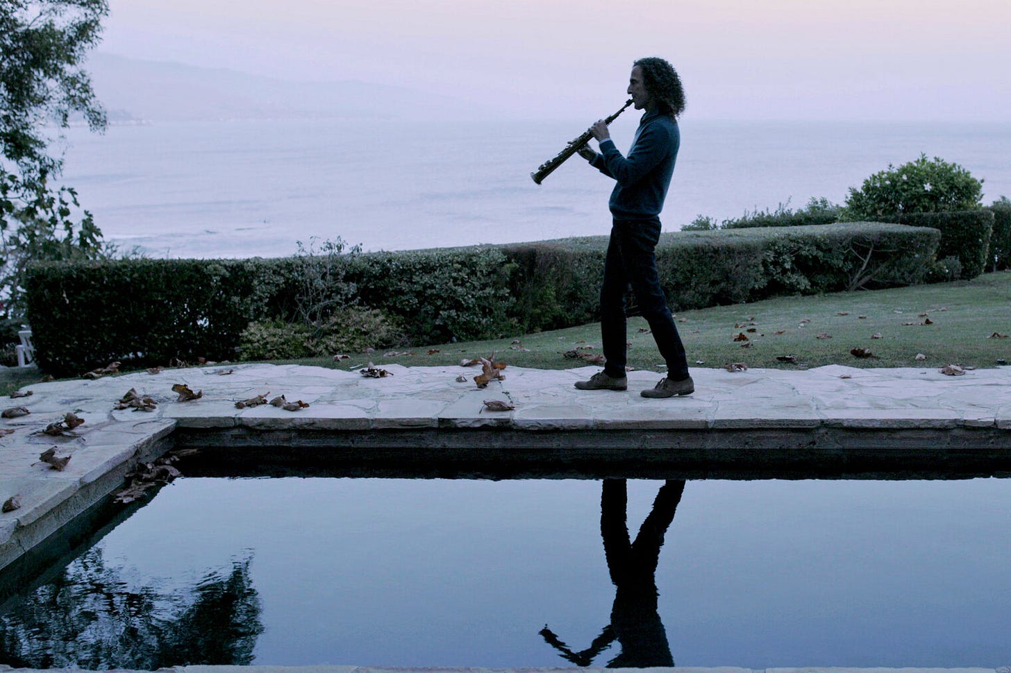 Kenny G blowing a sax by a pool; you can see his reflection