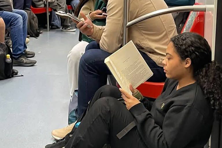 A imagem mostra um interior de metrô com várias pessoas sentadas e em pé. Uma mulher está sentada no chão, lendo um livro, enquanto um homem ao lado dela está olhando para o celular. Outros passageiros estão visíveis ao fundo, alguns também usando dispositivos móveis. O ambiente é típico de um transporte público, com assentos vermelhos e um piso azul.