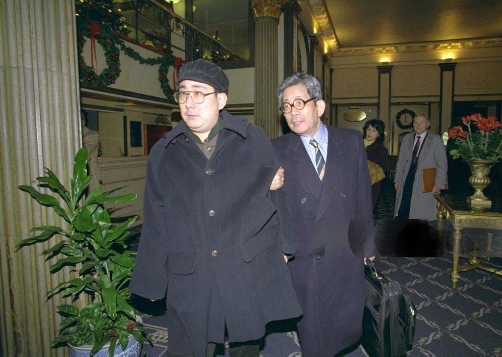 La imagen muestra a dos hombres mayores vestidos con abrigos oscuros y gafas, caminando en lo que parece ser el interior de un edificio ornamentado con decoraciones navideñas, como guirnaldas y flores. Detrás de ellos se pueden ver otras personas, posiblemente asistentes o invitados. El ambiente general sugiere que la imagen fue tomada en un entorno formal o institucional.