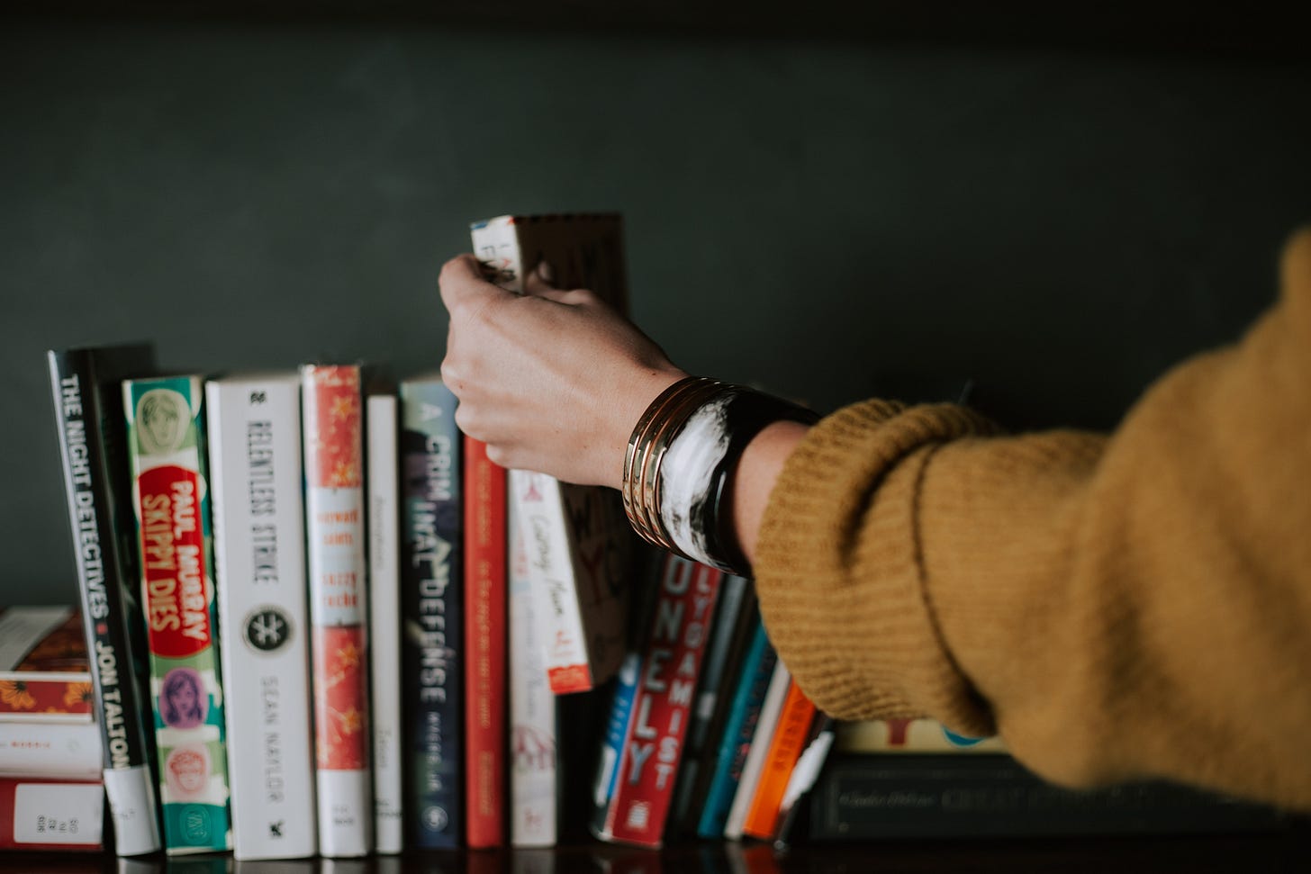 A hand reaches to choose a paperback book off a shelf of books.