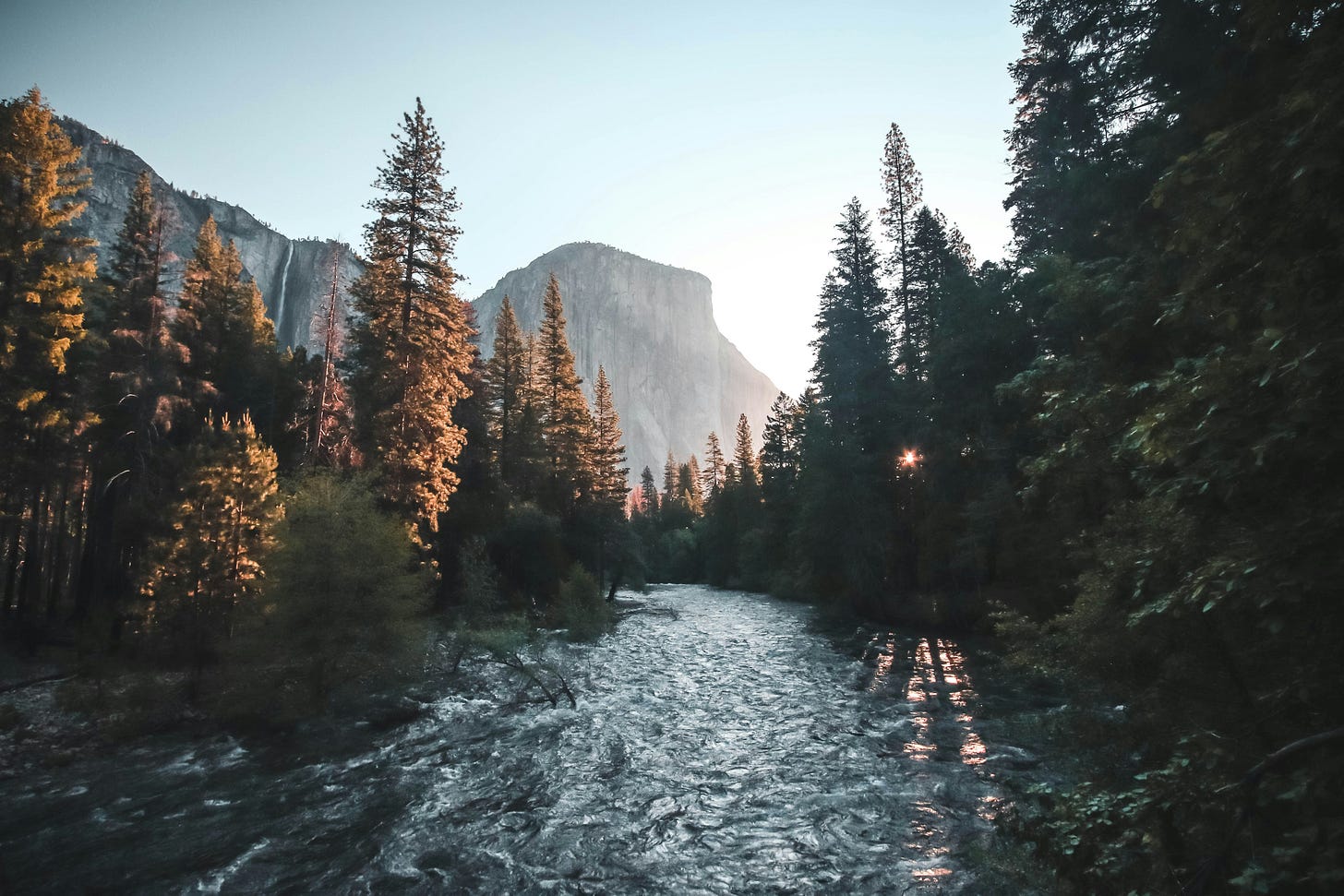 A dark river in low hazy sunlight, lined by pine trees. In the distance straight ahead is El Capitan. A dark river in low hazy sunlight, lined by pine trees. In the distance straight ahead is El Capitan.