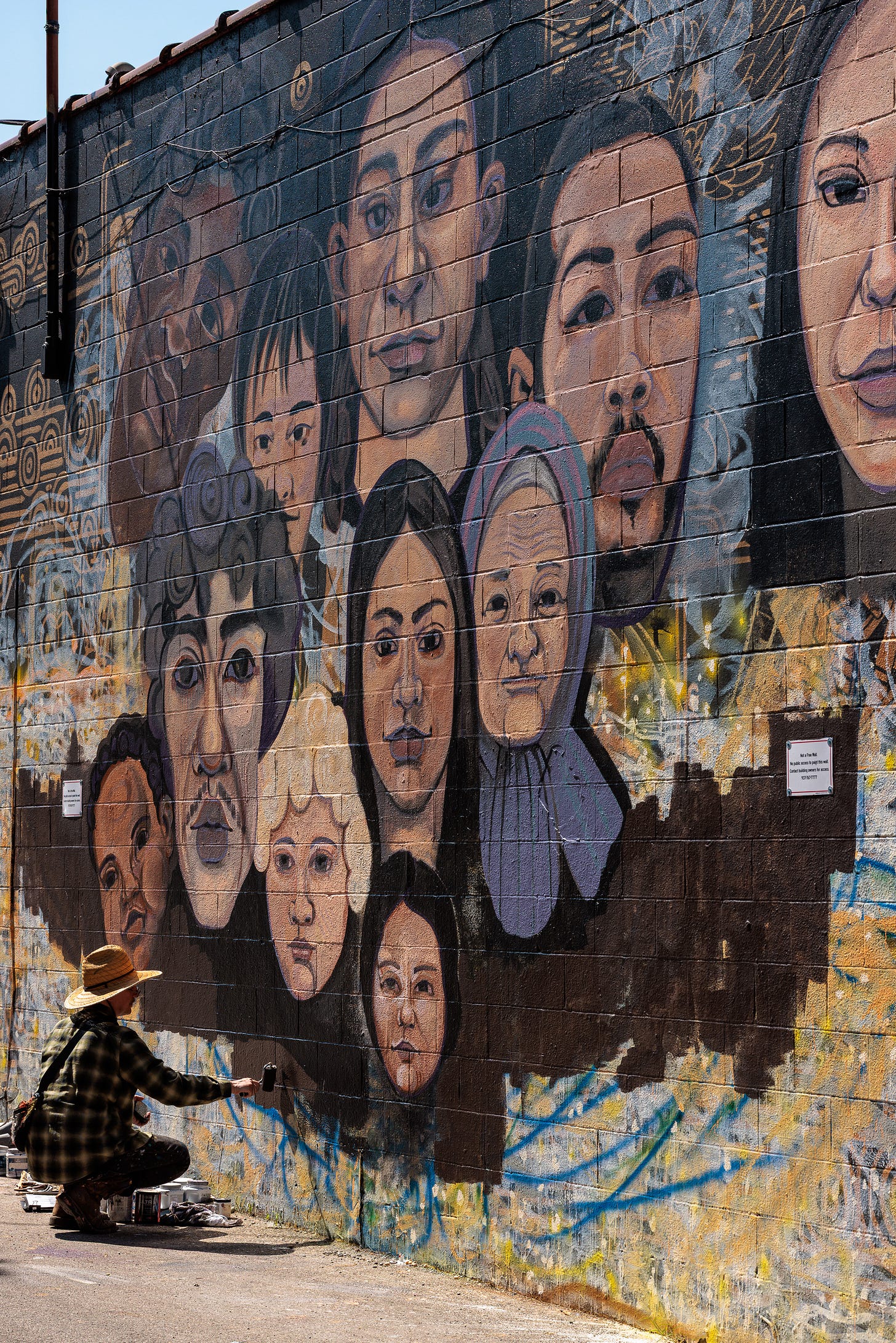 Pierre Nagley, wearing a straw hat, crouches on the ground painting the lower row of portrait faces on the mural, brush in hand.