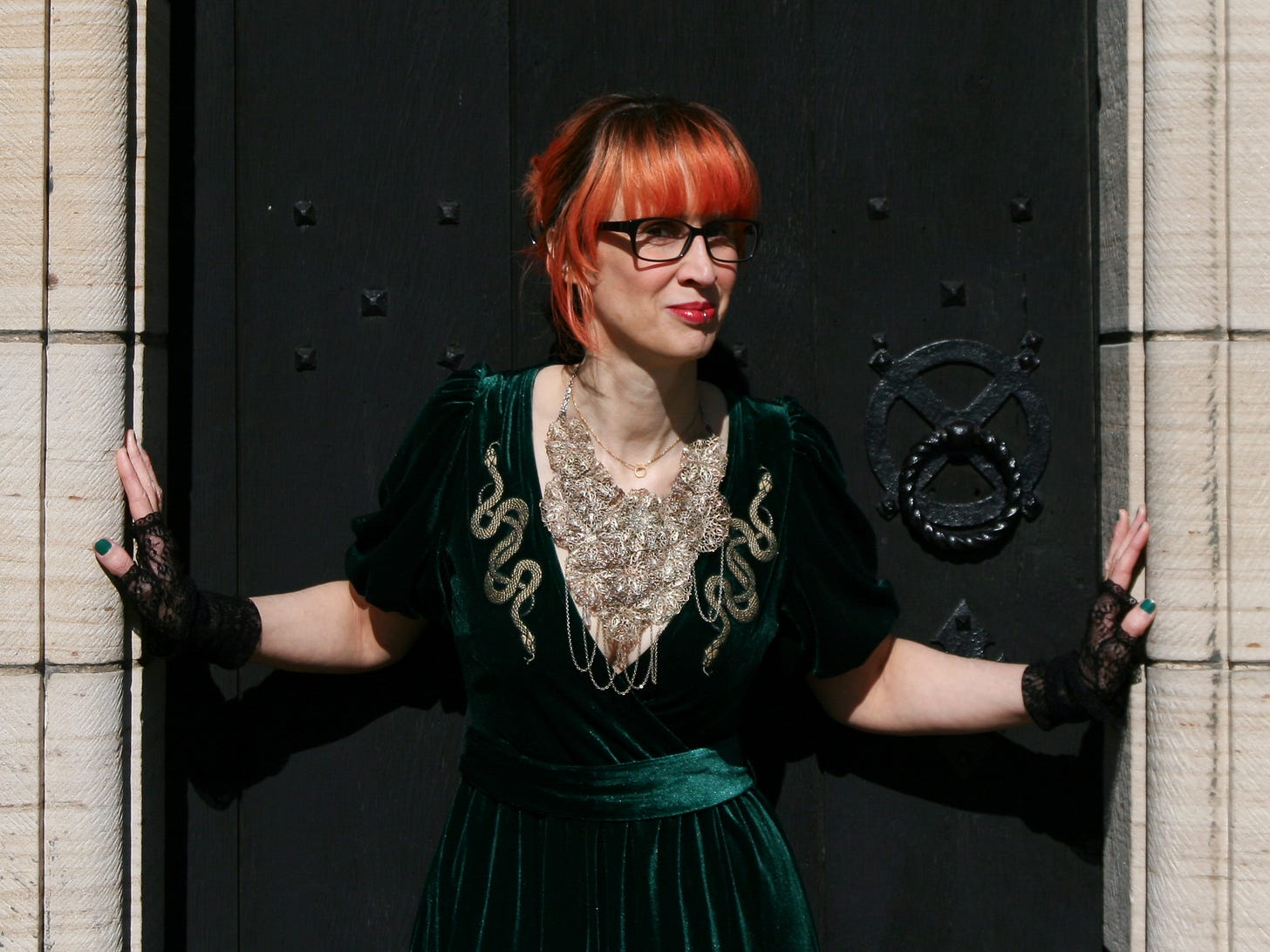 A thin white woman is standing in front of a black door, with her lace-gloved hands pressed against the doorway either side. She has bright orange hair pinned up and a long straight fringe, black-frame glasses, red lipstick, and a silver lip ring. Her head is partially turned to the right, but she’s looking to camera with a slight mischievous smile. She’s wearing a short-sleeved green velvet dress with two golden snake motifs running down the side from shoulder to breast. There’s a small golden apple necklace at her throat and a huge golden mesh necklace that covers her chest.