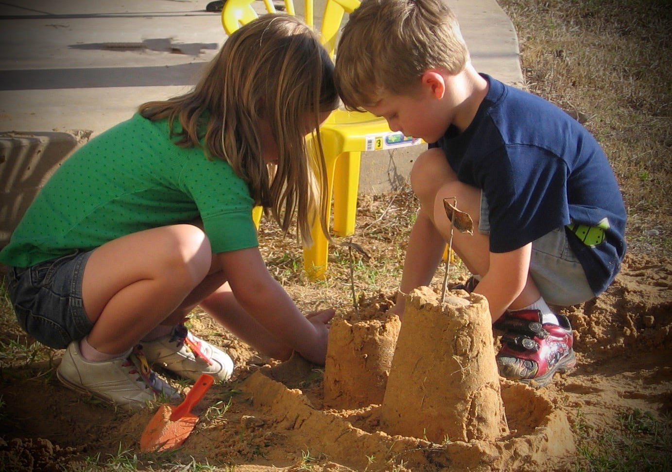 A young girl in a green shirt and boy in a blue shirt building sandcastles in the backyard