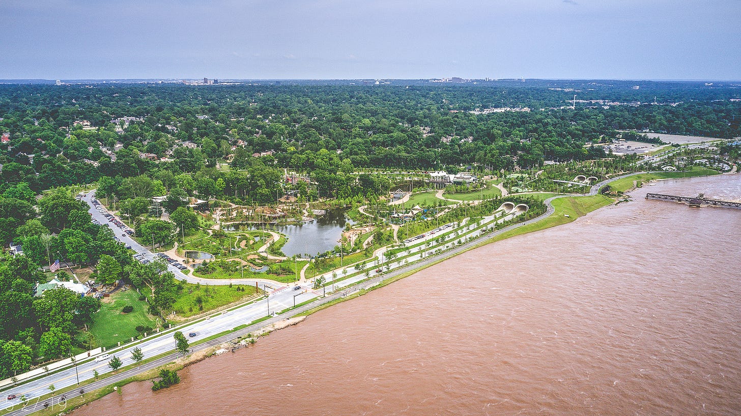 Seen from above, the muddy waters of the flooded Arkansas River reach almost to the River Parks trail at Gathering Place: a large park with carnival-like structures, an irregularly shaped pond, and winding paths around extensive landscaping. The backdrop is a lush, expansive view of midtown and south Tulsa below a slightly hazy, cornflower blue sky. Inside Gathering Place, the parking lot appears full. Seen from above, the muddy waters of the flooded Arkansas River reach almost to the River Parks trail at Gathering Place: a large park with carnival-like structures, an irregularly shaped pond, and winding paths around extensive landscaping. The backdrop is a lush, expansive view of midtown and south Tulsa below a slightly hazy, cornflower blue sky. Inside Gathering Place, the parking lot appears full.