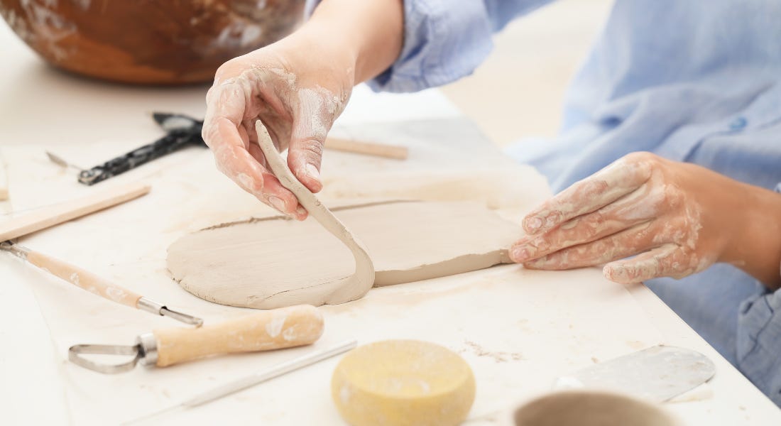 A pair of hands working on a grey slab of clay, with tools on the table around the white table A pair of hands working on a grey slab of clay, with tools on the table around the white table