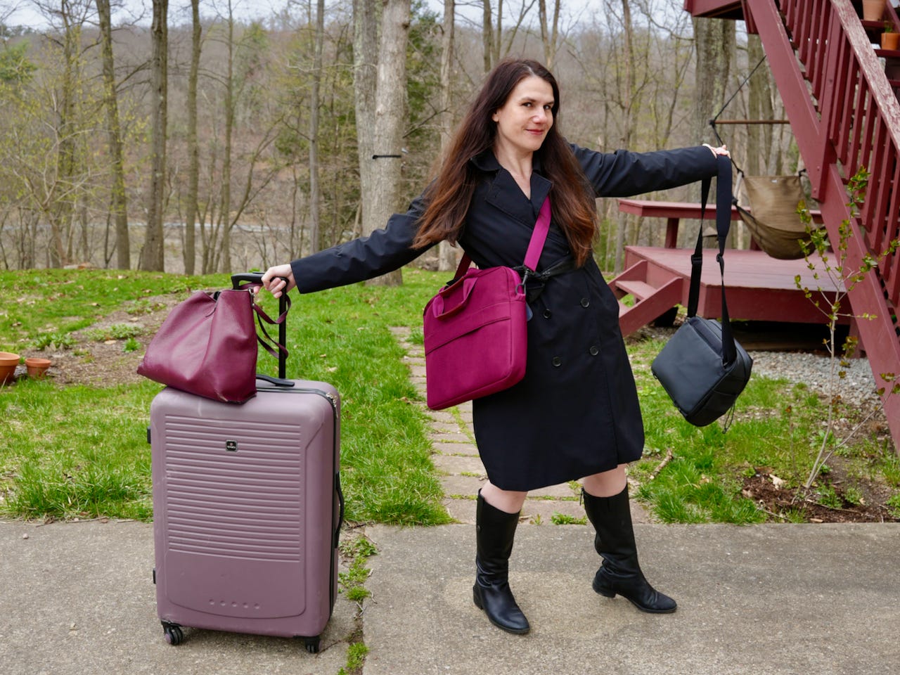 A woman (Alexandra Hidalgo) stands outside. She has a large suitcase and a purse, there’s a computer bag over her shoulder. On her other hand there’s a camera bag. She’s smiling at the camera. There is grass and trees behind her. It’s afternoon.