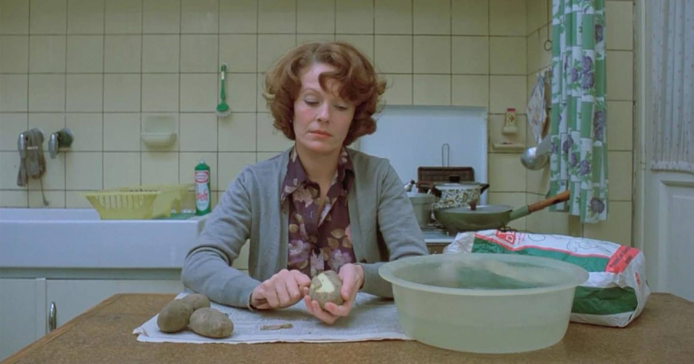 Still from Jeanne Dielman (1975), showing Delphine Seyrig seated in a sterile kitchen, peeling potatoes at a brown laminate counter—an iconic image of domestic repetition and cinematic minimalism.