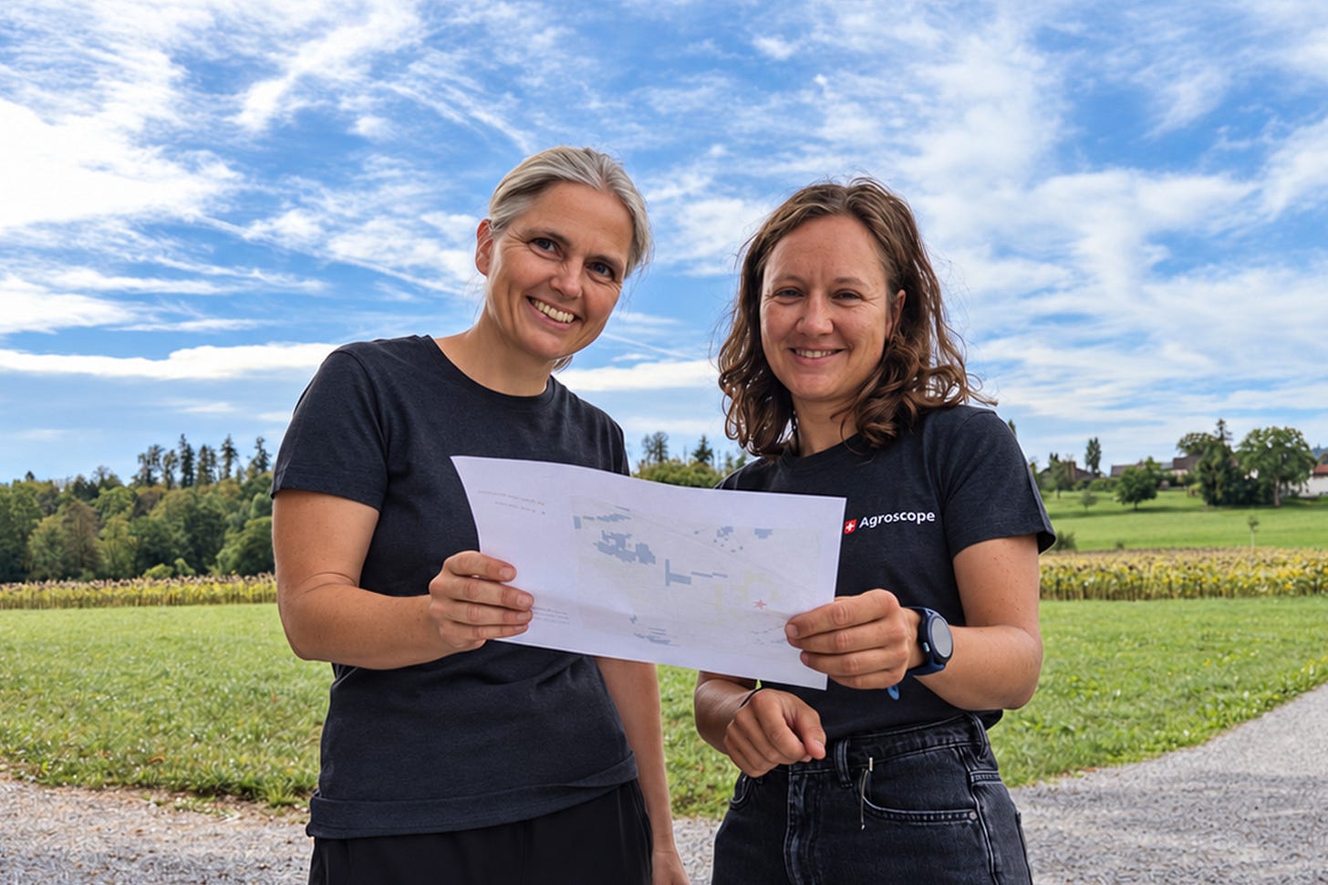 Die Forstwirtschaftlerin und Geographin Sonja Kay (links) und die Geographin Amelie Kreuzer (rechts) stehen lächelnd auf einem Feldweg und halten gemeinsam einen ausgedruckten Plan in die Kamera. Beide tragen dunkle T-Shirts, Kreuzer eines mit dem Agroscope-Logo. Im Hintergrund erstrecken sich grüne Wiesen, ein Maisfeld und eine leicht hügelige Landschaft unter blauem, wolkigem Himmel. Die Forstwirtschaftlerin und Geographin Sonja Kay (links) und die Geographin Amelie Kreuzer (rechts) stehen lächelnd auf einem Feldweg und halten gemeinsam einen ausgedruckten Plan in die Kamera. Beide tragen dunkle T-Shirts, Kreuzer eines mit dem Agroscope-Logo. Im Hintergrund erstrecken sich grüne Wiesen, ein Maisfeld und eine leicht hügelige Landschaft unter blauem, wolkigem Himmel.