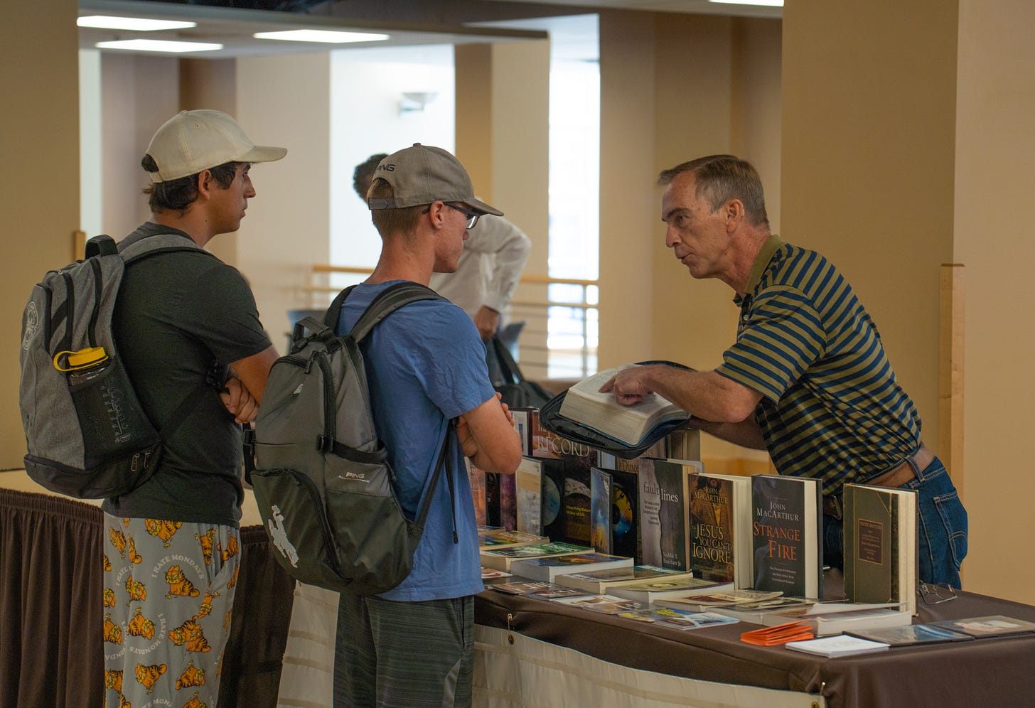 An older man gestures to a line in a bible as two students in ballcaps look on, arms folded. An older man gestures to a line in a bible as two students in ballcaps look on, arms folded.