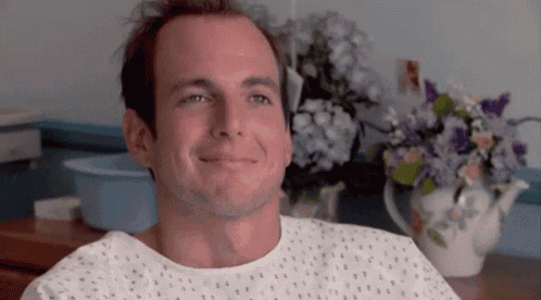 a man in a hospital gown is smiling in front of a vase of flowers .