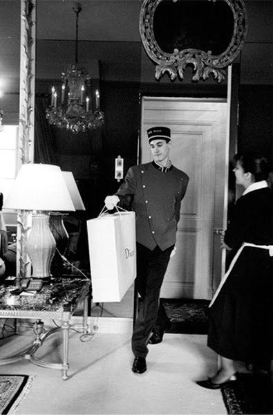This may contain: a black and white photo of a man in uniform holding shopping bags while standing in front of a chandelier
