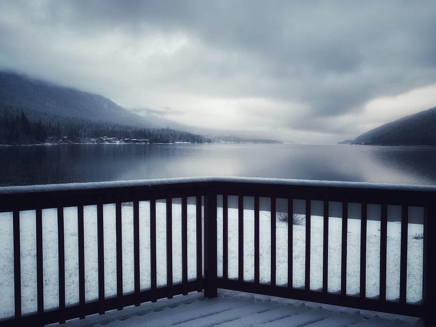 Snow-covered deck railing overlooking Kootenay Lake in Nelson, British Columbia, with calm water, forested mountains, and a cloudy winter sky.