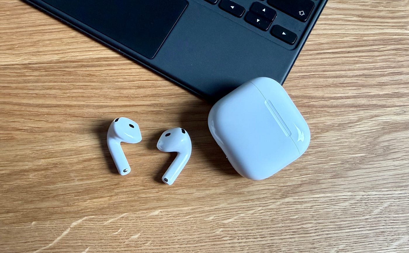 Apple AirPods wireless earbuds with charging case placed next to iPad keyboard on wooden desk.