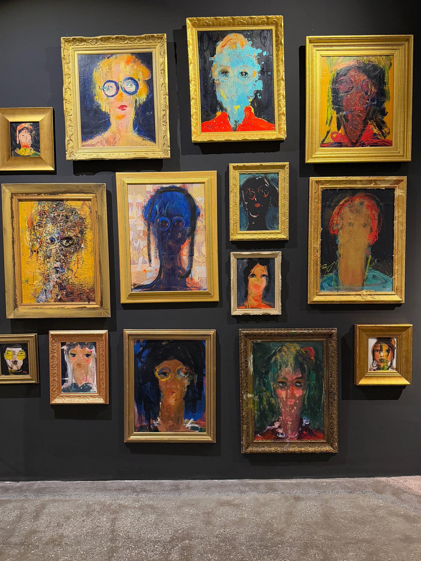 A gallery wall with multiple framed paintings by Habib Kibari from the Aliens series. The works show distorted, abstract human faces with smeared colors, obscured features, and expressive brushstrokes, arranged salon-style in ornate gold frames against a dark wall. A gallery wall with multiple framed paintings by Habib Kibari from the Aliens series. The works show distorted, abstract human faces with smeared colors, obscured features, and expressive brushstrokes, arranged salon-style in ornate gold frames against a dark wall.