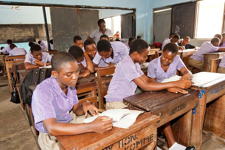 A classroom full of students in uniform reading from textbooks while a teacher looks on A classroom full of students in uniform reading from textbooks while a teacher looks on