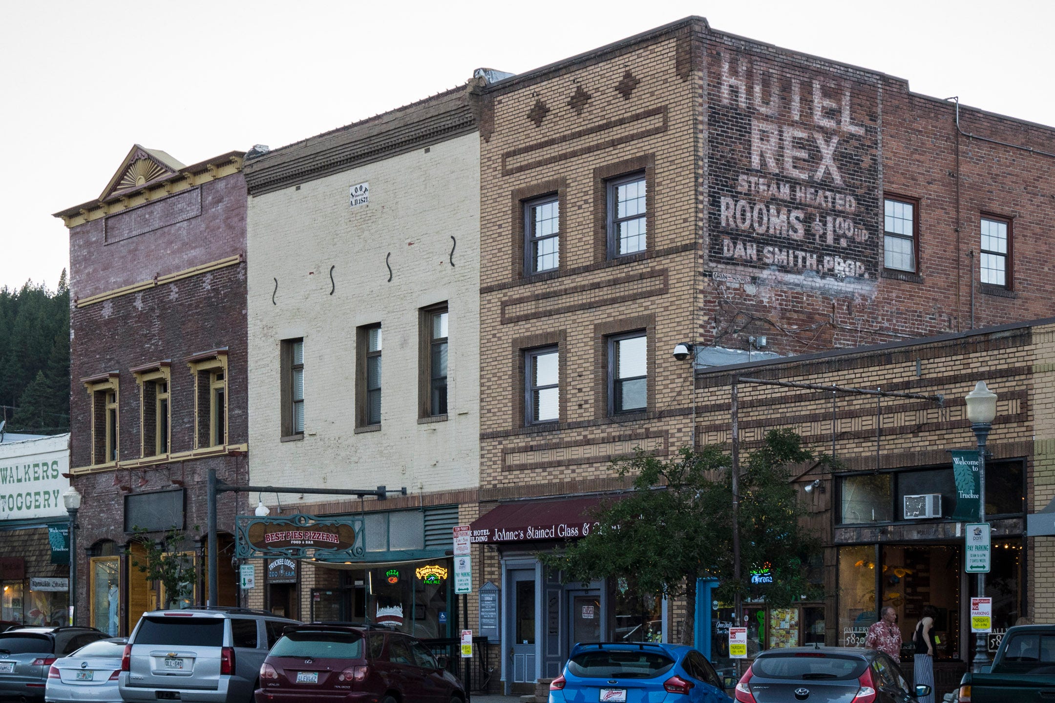 A row of old, brick buildings in Truckee, California. The main grouping of buildings are three stories, with retail on the first floor and rooms above. One building has a pizzeria. The other a stained glass shop with a maroon awning. Each building has a simple and distinctive cornice at the top. One of the buildings has a faded, old advertisement painted on the brick side, "HOTEL REX STEAM HEATED ROOMS $1.00 UP. DAN SMITH, PROP." Each building is in a different state up upkeep/disrepair. Smaller, 1 story building flank the row of three. Cars are parked on the curb and a pair of pedestrians are window shopping.