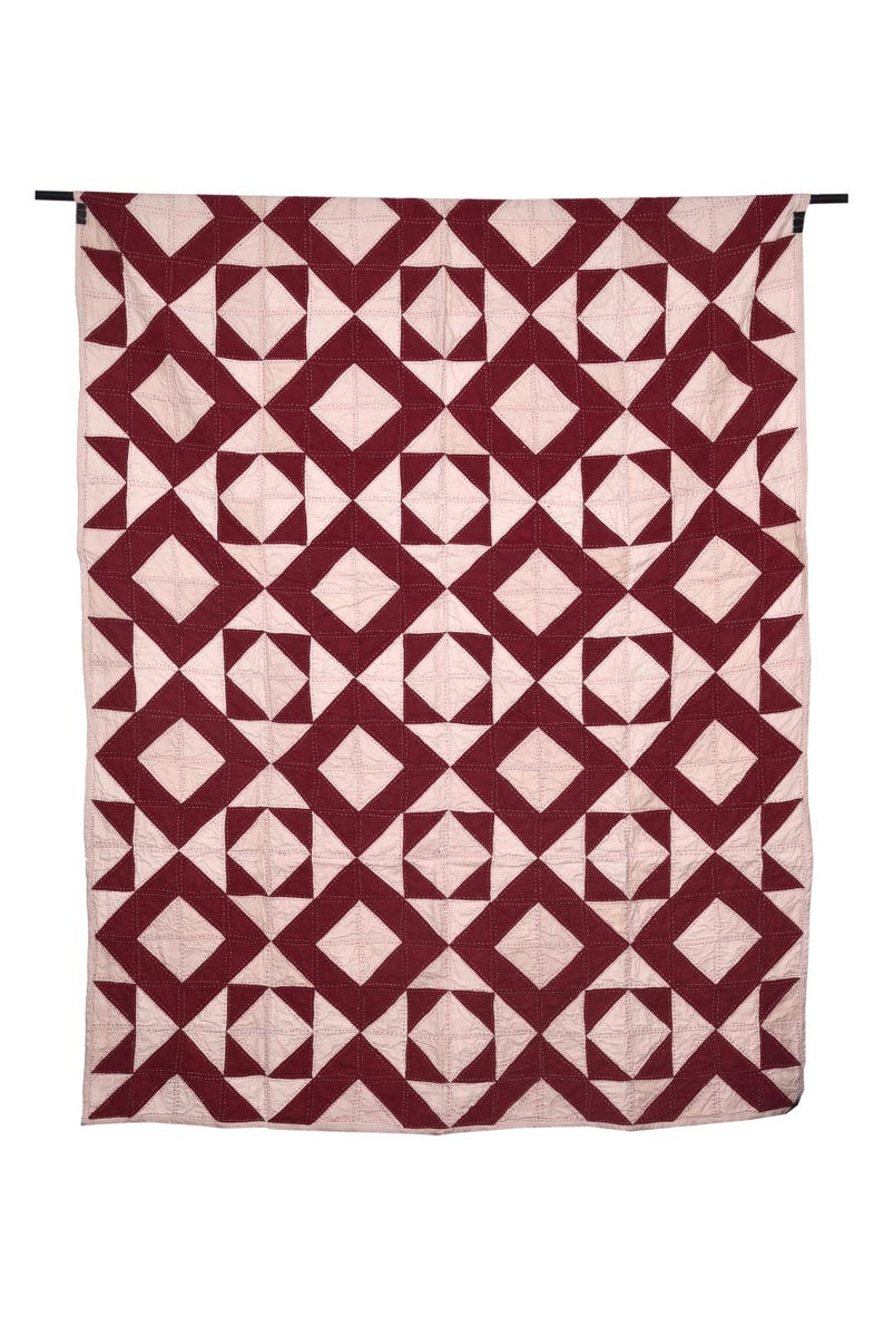 May include: A handmade quilt with a geometric pattern. The quilt features a repeating design of burgundy diamonds and triangles set against a light pink background. The quilt is hanging from a black rod. May include: A handmade quilt with a geometric pattern. The quilt features a repeating design of burgundy diamonds and triangles set against a light pink background. The quilt is hanging from a black rod.