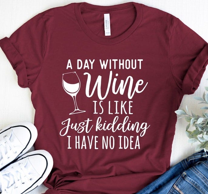 May include: A maroon t-shirt with white text that reads 'A day without wine is like just kidding I have no idea'. The text is surrounded by a white outline of a wine glass. The t-shirt is laid out on a white surface with a pair of white sneakers and a pair of blue jeans.
