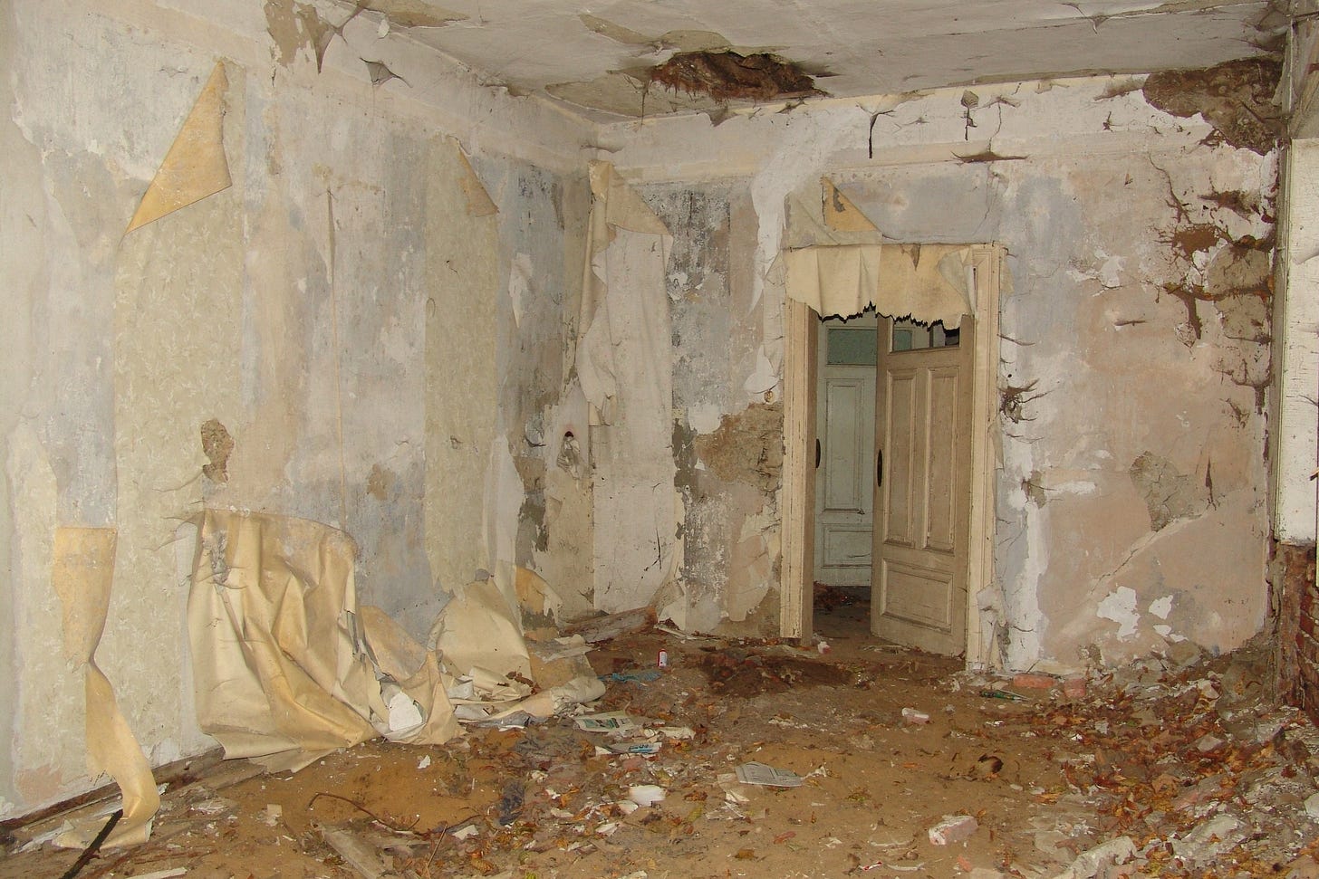 A room with peeling wallpaper, a hole in the ceiling, and crumbling brickwork