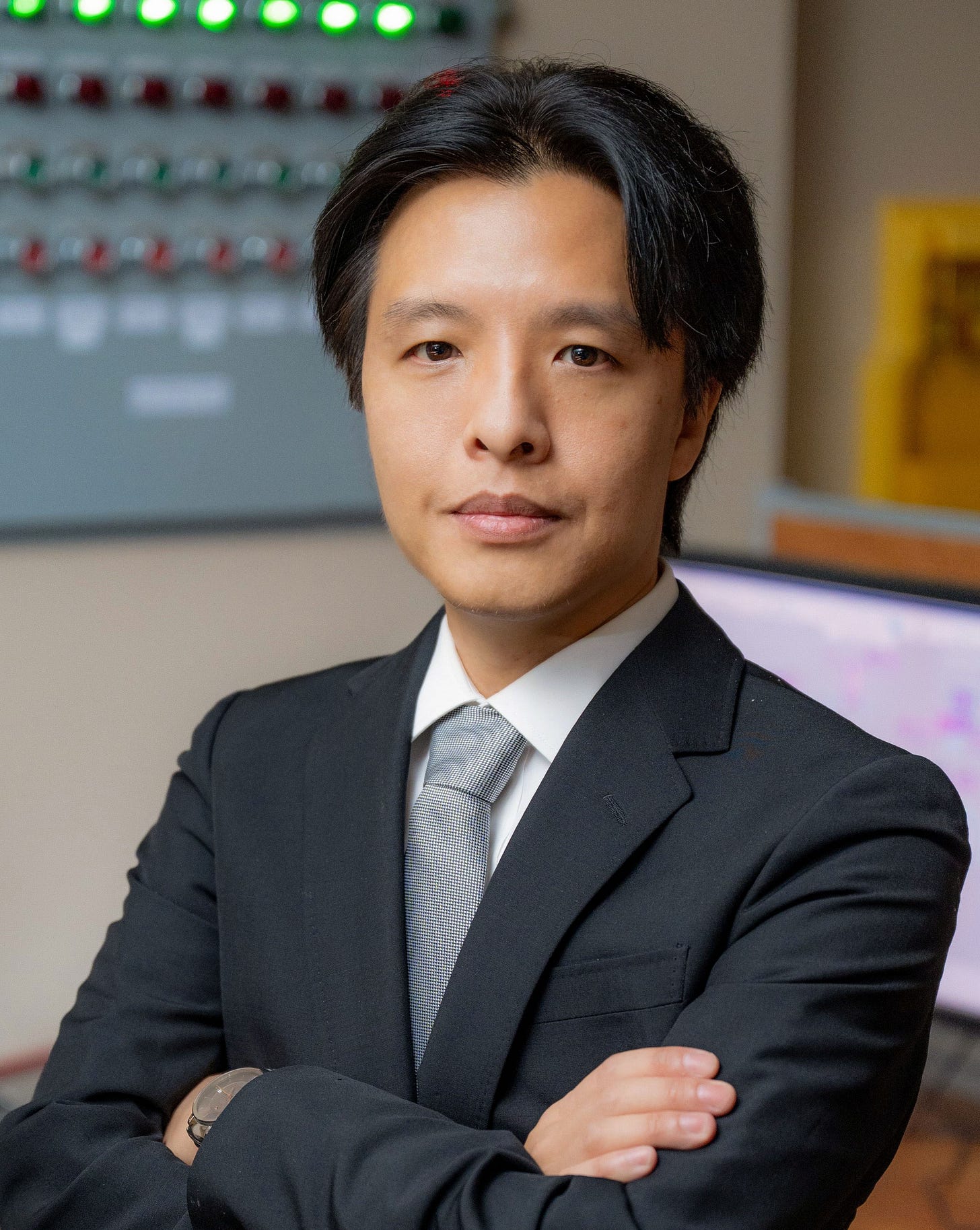 Roger Chin, Software Architect at Applied Atomics, in a dark suit and gray tie, arms crossed, standing in front of an indicator light panel and monitor in the IST control room.