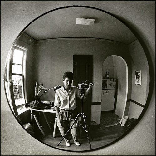 A black-and-white photo shows Lola reflecting in a round mirror, standing with a camera tripod in a cozy room. The scene feels introspective and artistic. There are windows to the right of them, and an arched doorway to the left of them. Lola has short hair and is staring straight into their reflection. A black-and-white photo shows Lola reflecting in a round mirror, standing with a camera tripod in a cozy room. The scene feels introspective and artistic. There are windows to the right of them, and an arched doorway to the left of them. Lola has short hair and is staring straight into their reflection.