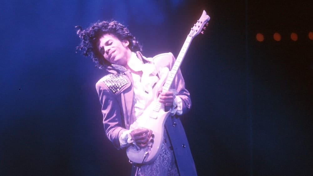 Prince, bathed in purple light and wearing a flamboyant outfit, plays his guitar in a scene from Purple Rain