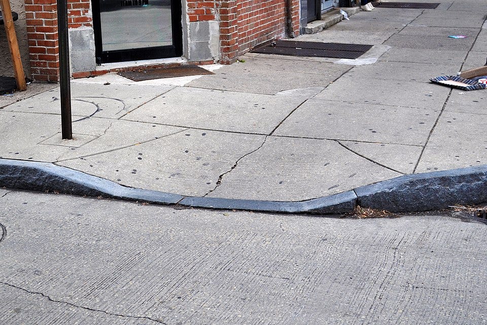 A weathered curb cut on a cracked concrete sidewalk next to a brick building with a large window. The curb cut, designed for accessibility, shows signs of wear with uneven edges and surrounding pavement fissures. Debris, including a small pile of dirt and a piece of cloth, is scattered nearby. A metal pole and a grate are visible near the building entrance. Photo credit: Nick-philly