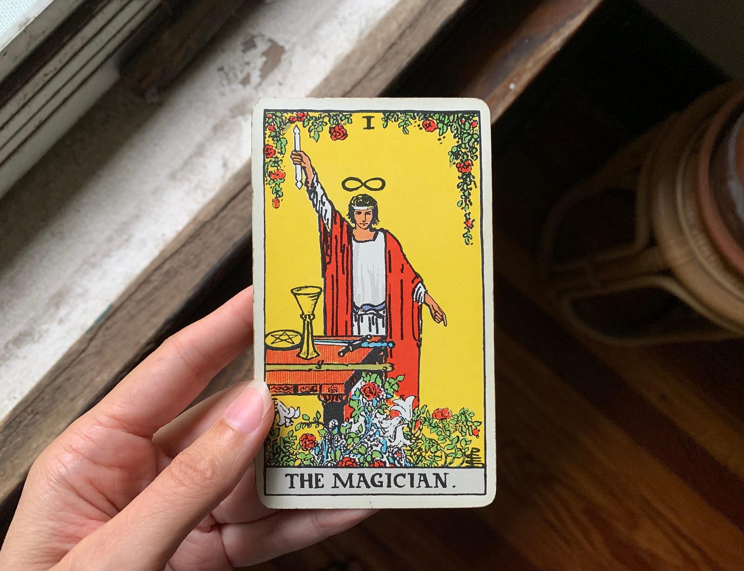 A hand is holding a tarot card, The Magician by Pamela Colman Smith for the Rider Waite Smith Tarot. In the image a person is wearing a white robe and a red vest a an ouroboros belt. They have an infinity symbol above their head, a wand in one hand pointed up to the sky and are pointing the other hand down toward the earth. There's a desk in front of them with four items on it, a pentacle, cup, sword and wand.