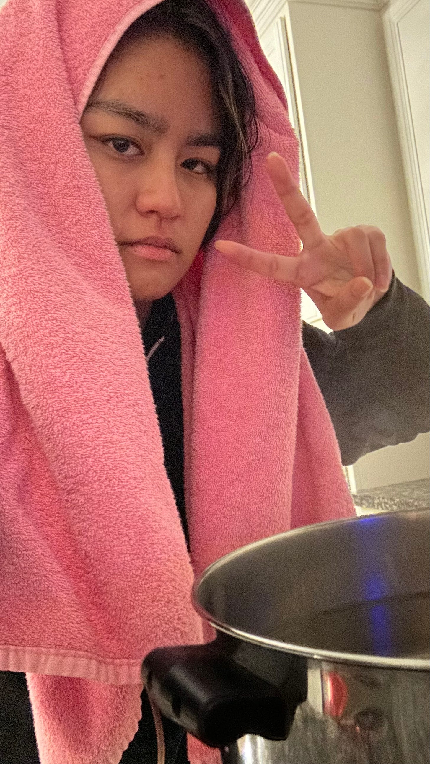 Joelle taking a selfie in front of a steaming pot on a stove with a pink towel draped over her head. She makes a blank facial expression and is holding up a peace sign with her free hand