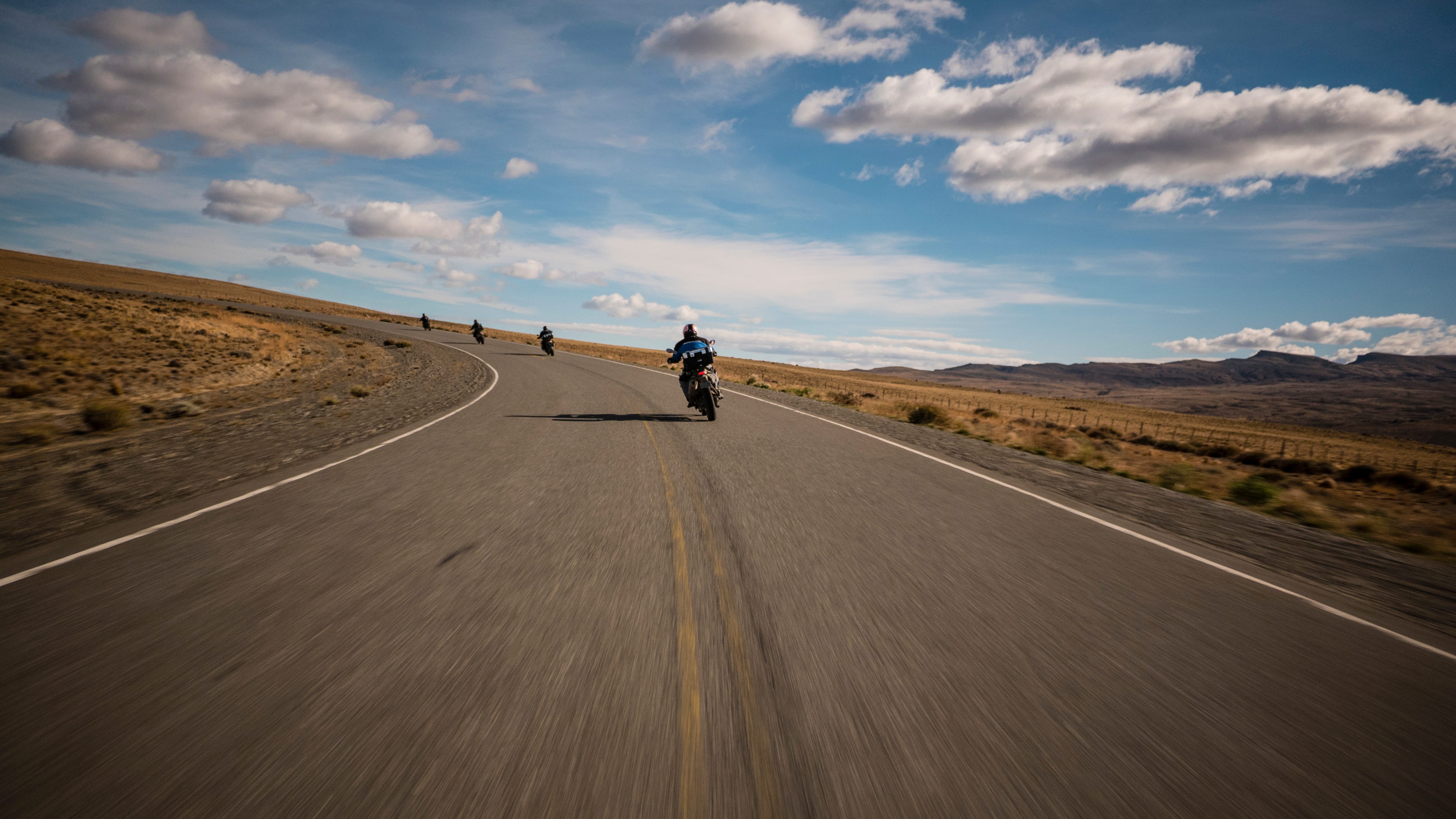 Photo of a road bending left across a broad, barren landscape. The point of view if from a motorcycle bending into the turn, with four motorcycles in front, also bending into the turn. The horizon is tilted and the foreground is blurred, creating a sense of speed. The sky is blue and dotted with clouds. On the right horizon are hills. The road continues to the left.