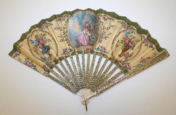 a hand fan with elaboration floral decorations and a picture of two women sitting and a man standing above them in the center