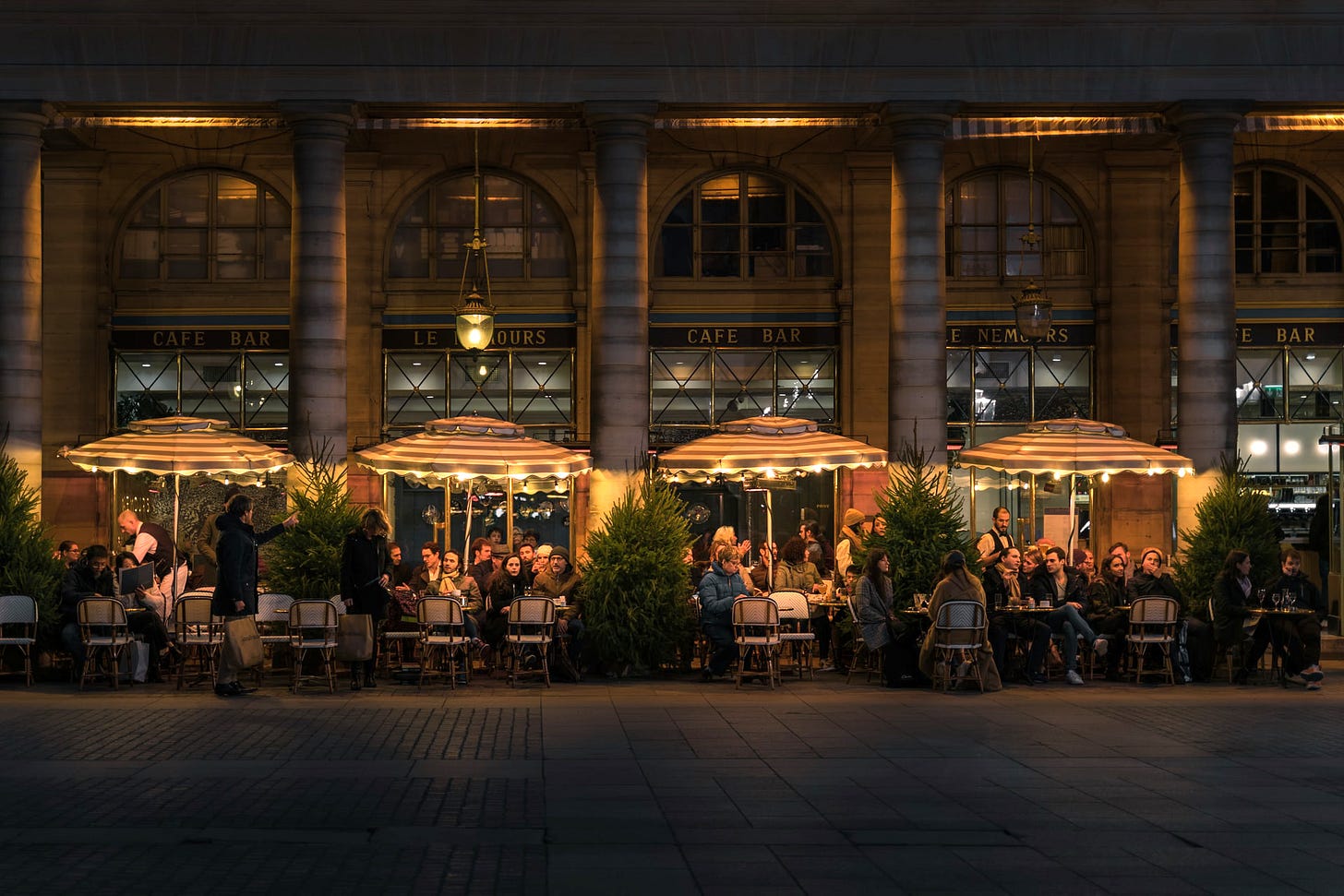 A photo of a Parisian street cafe at night. Many people are seated outside, under illuminated umbrellas.