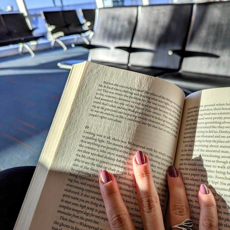 Photo of an airport departure hall, chairs brightly lit by sunlight. A book is open, the sun illuminated a portion of the page, and a hand holds it open.