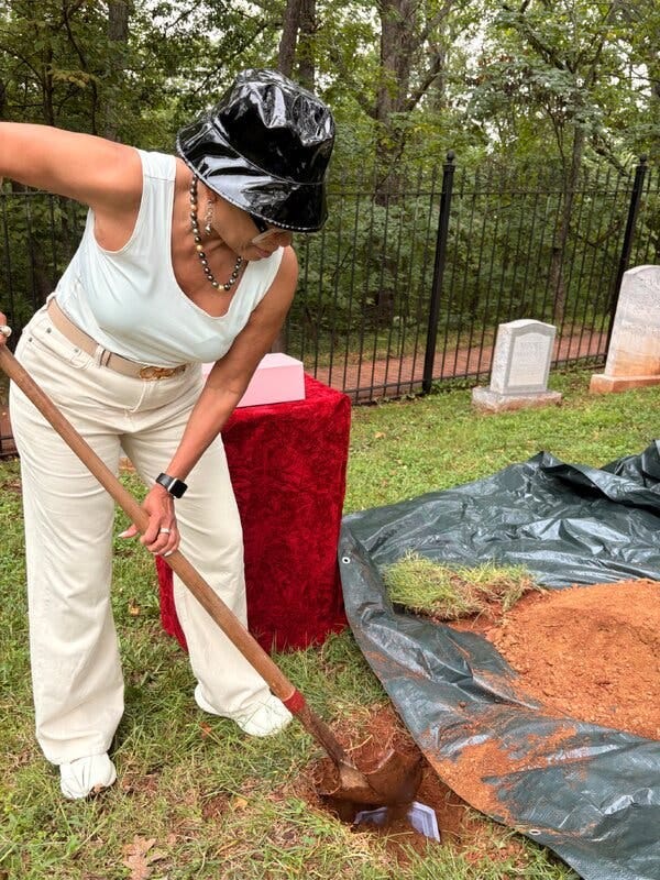 A woman wearing white pants and shirt digs into the ground with a shovel. Gravestones are lined up next to a fence.