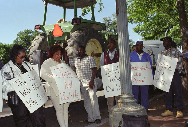color photo of protesters standing in front of a green farm tractor on a trailer, carrying signs that say 'Justice Not Just-us,' 'Don't take our land,' 'Discrimination is illegal' and 'The Lord will make a way somehow'