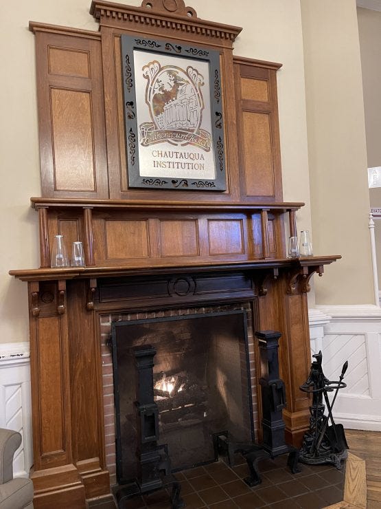 An elegant Victorian-era fireplace with a tall wooden mantel and overmantel featuring a framed Chautauqua Institution logo. The mantel includes decorative moldings, a display shelf with glass vases, and is flanked by black cast iron andirons. A fire burns in the brick-lined firebox, and white wainscoting is visible on the adjacent wall.