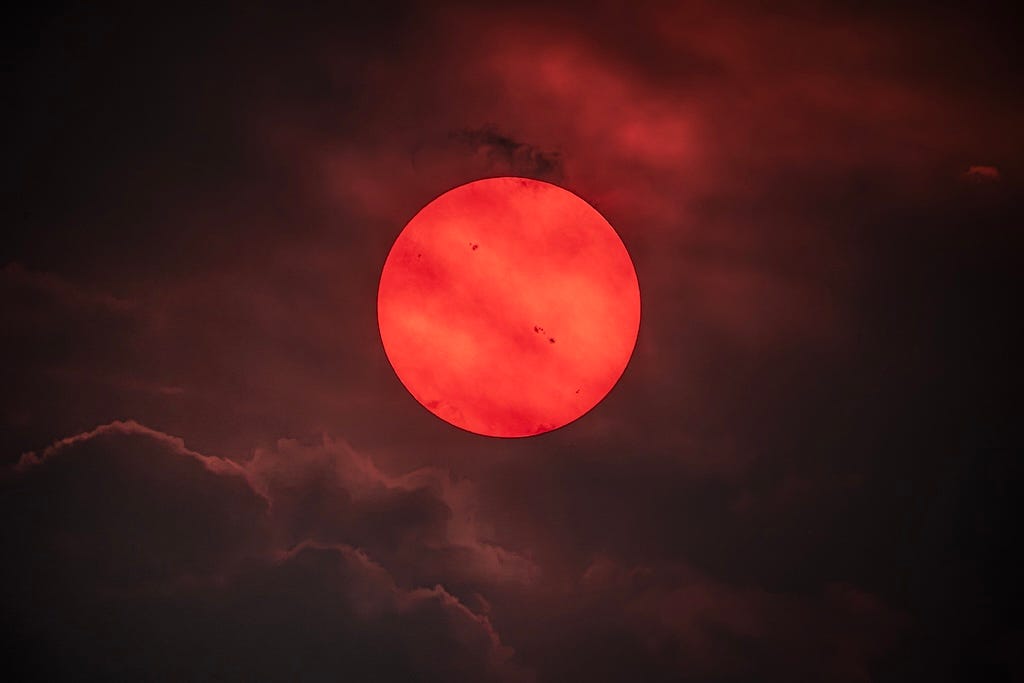 A deep red sun shows several sunspots.
Clouds and haze are visible. A deep red sun shows several sunspots.
Clouds and haze are visible.