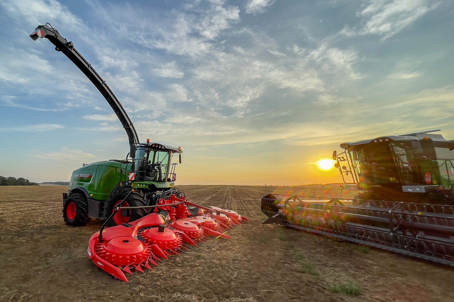 Zwei grosse landwirtschaftliche Maschinen der Marke Fendt stehen auf einem abgeernteten Feld bei Sonnenuntergang. Links ist ein grüner Feldhäcksler mit einem auffälligen roten Maisgebiss und einem langen, nach oben geschwungenen Auswurfarm zu sehen. Rechts daneben befindet sich ein Mähdrescher mit einem breiten Schneidwerk. Der Himmel ist leicht bewölkt, während die tief stehende Sonne die Szene in ein warmes Licht taucht. Zwei grosse landwirtschaftliche Maschinen der Marke Fendt stehen auf einem abgeernteten Feld bei Sonnenuntergang. Links ist ein grüner Feldhäcksler mit einem auffälligen roten Maisgebiss und einem langen, nach oben geschwungenen Auswurfarm zu sehen. Rechts daneben befindet sich ein Mähdrescher mit einem breiten Schneidwerk. Der Himmel ist leicht bewölkt, während die tief stehende Sonne die Szene in ein warmes Licht taucht.