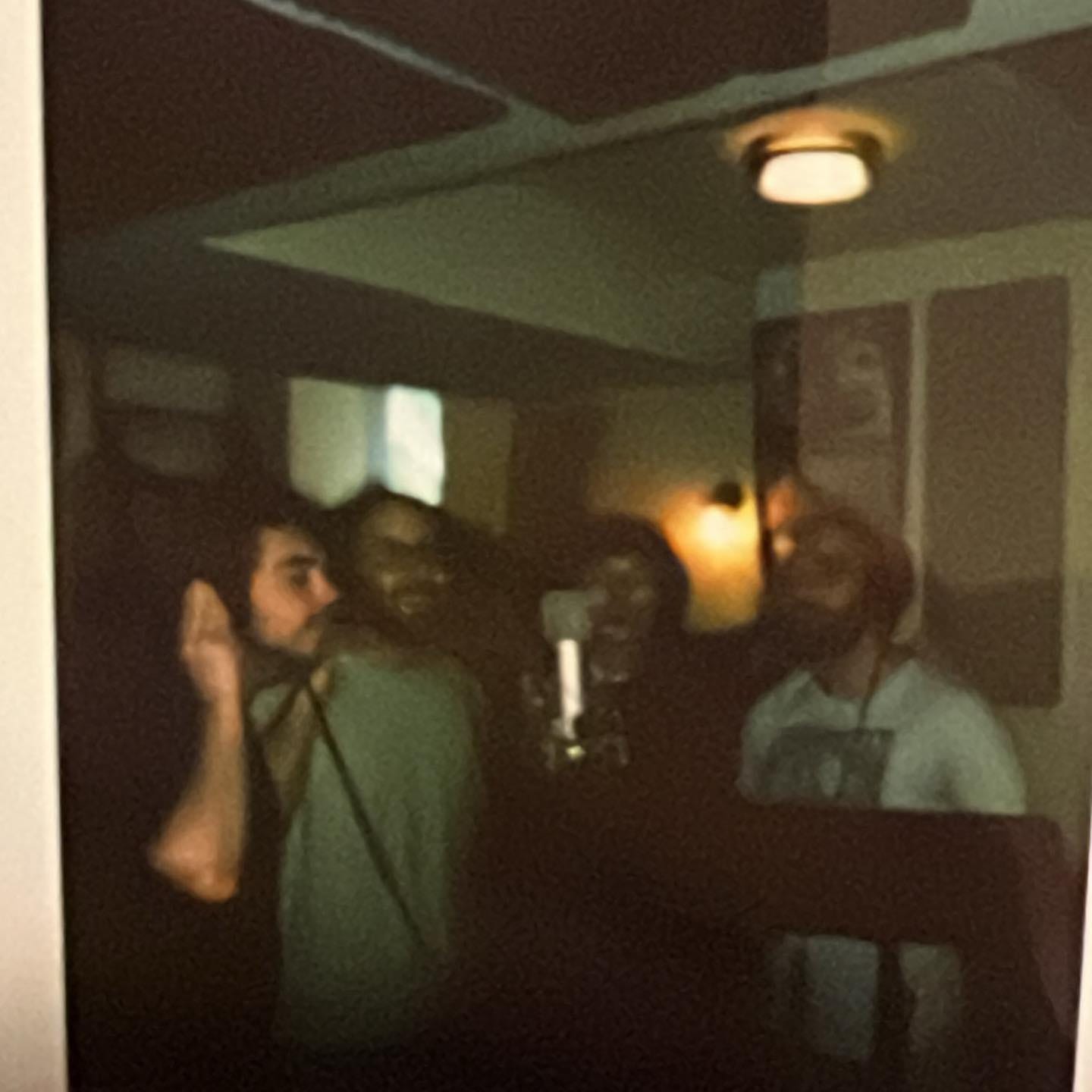 An underexposed Fujifilm photo of the band performing background vocals around a single microphone.