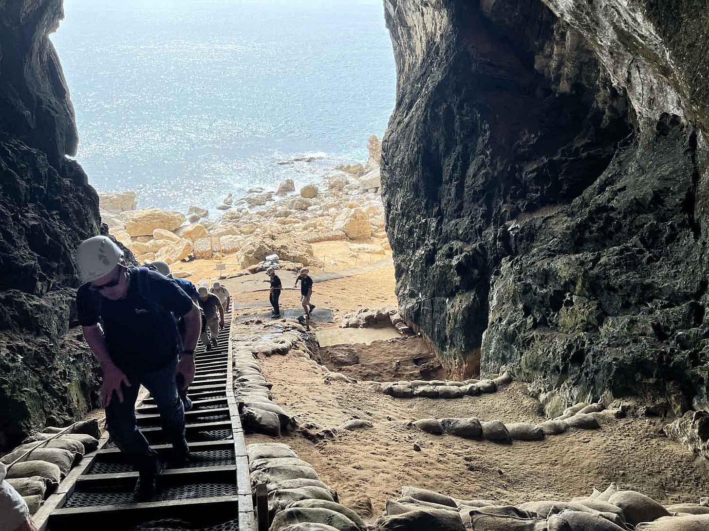 View from halfway up the interior slope of Vanguard Cave with a sloping pathway at left with people climbing upward, archaeological sandbagged deposits at right, and the sea visible outside the cave mouth View from halfway up the interior slope of Vanguard Cave with a sloping pathway at left with people climbing upward, archaeological sandbagged deposits at right, and the sea visible outside the cave mouth