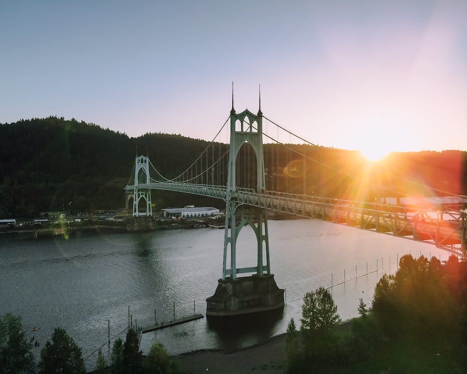 Free Magnificent scenery of aged St Johns Bridge crossing rippling river surrounded by hills with lush green woods against cloudless sunset sky in Portland Stock Photo
