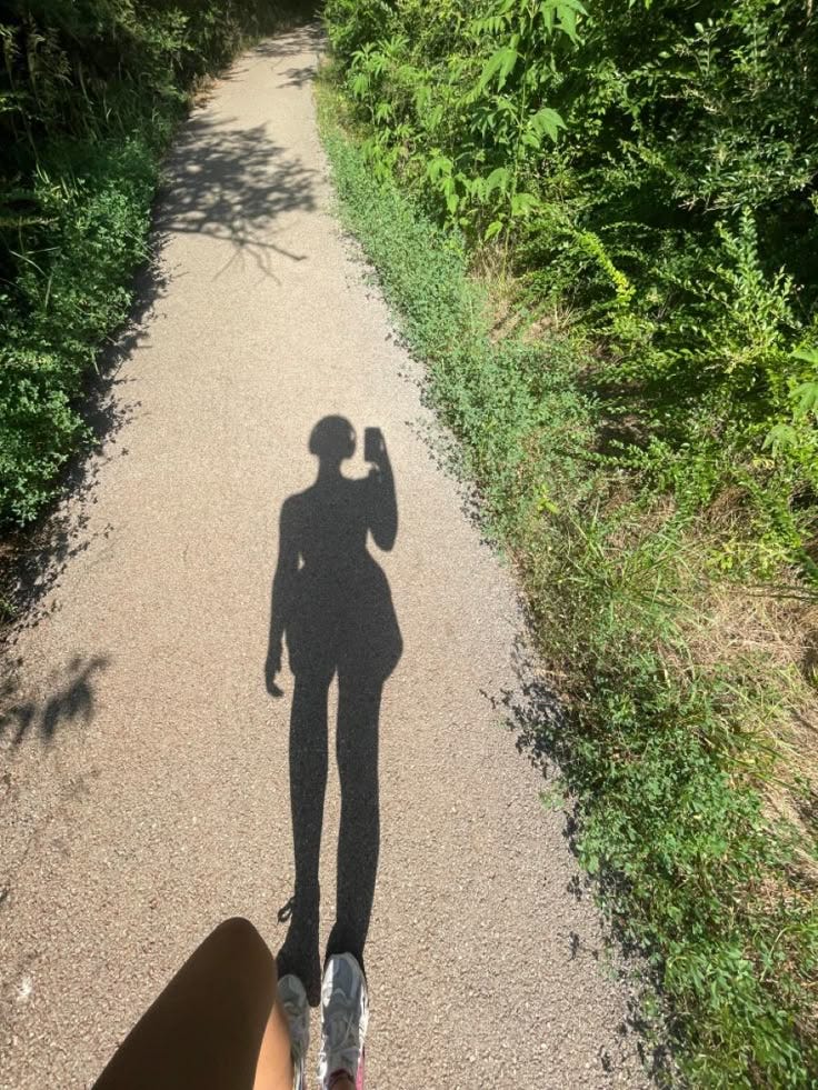 This may contain: the shadow of a person standing on a dirt road in front of bushes and trees