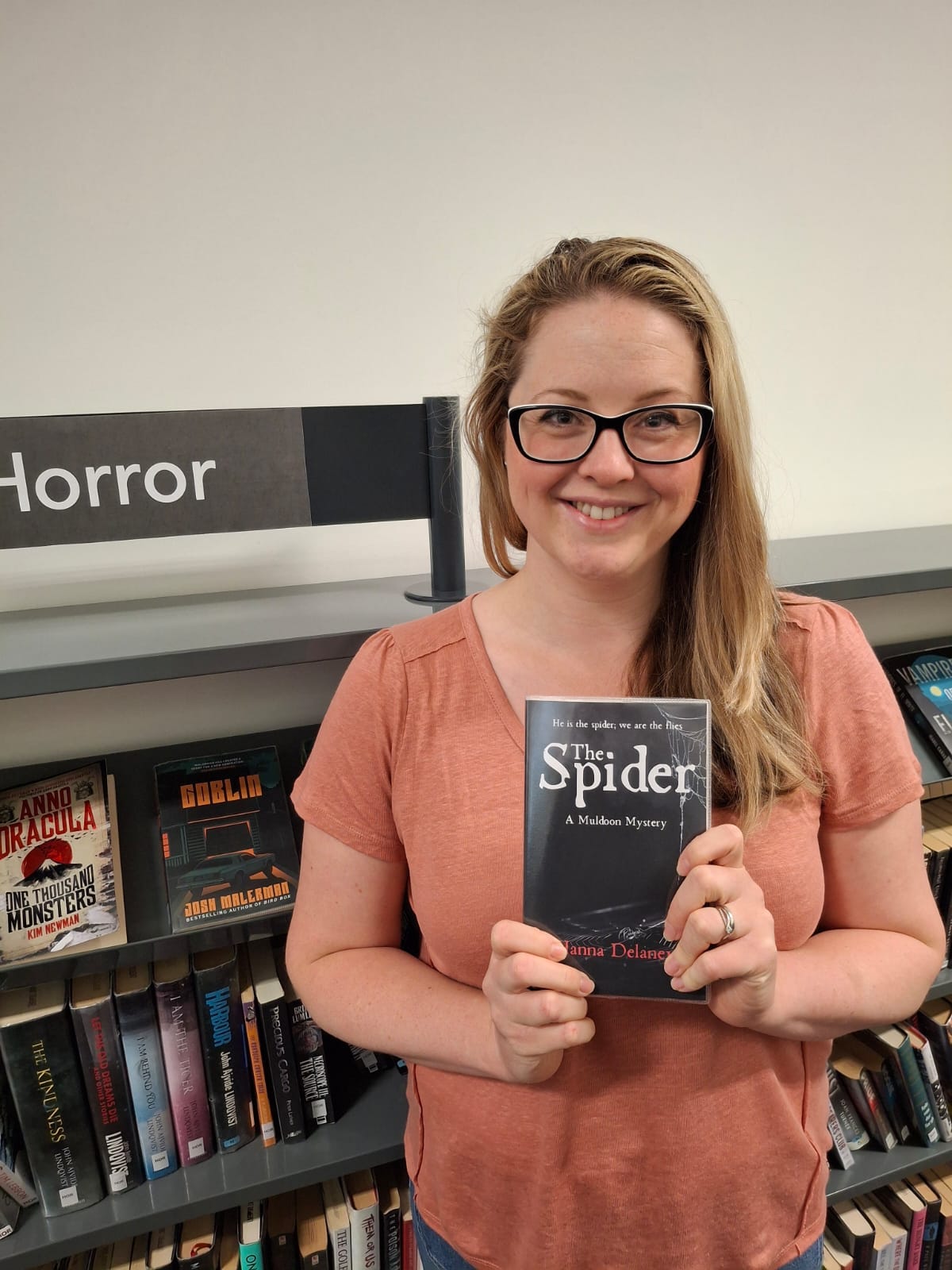 Hanna is standing in the horror section of the library holding a copy of her book, The Spider. The Spider is a gothic horror novel set in Victorian Liverpool. The photo was taken at Liverpool Central Library.